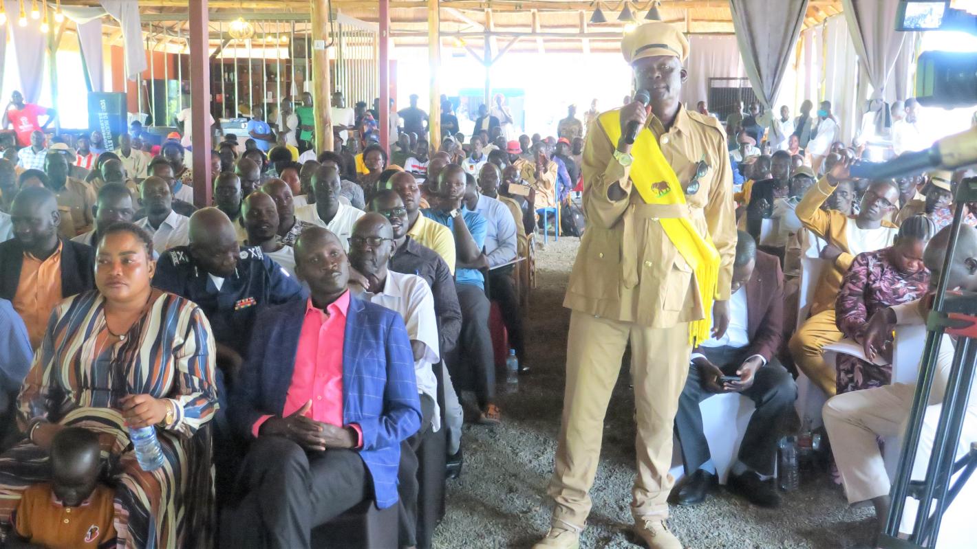 A military person standing in a yellow uniform wearing a yellow sash is speaking into a microphone to a large room of people seated in several rows behind them.