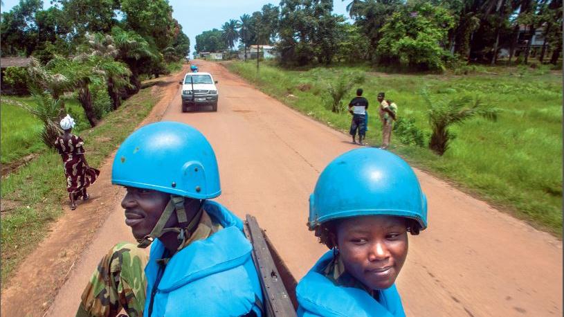 Peacekeepers sit on the back of a truck, travelling down a dirt road