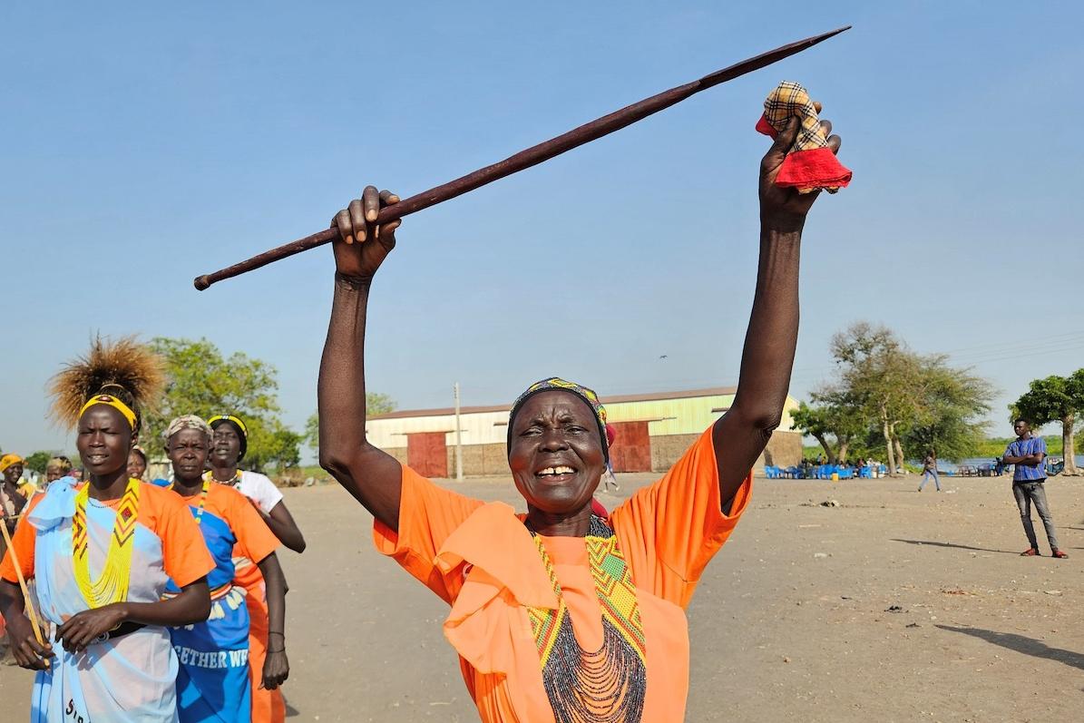There were happy faces all around when residents of Kodok in Upper Nile State celebrated International Women's Day, advocating for equal rights and empowerment. A woman wearing a bright orange t-shirt and large necklace is smiling and holding a wooden object above their head in jubilation. There are some other women in the background.