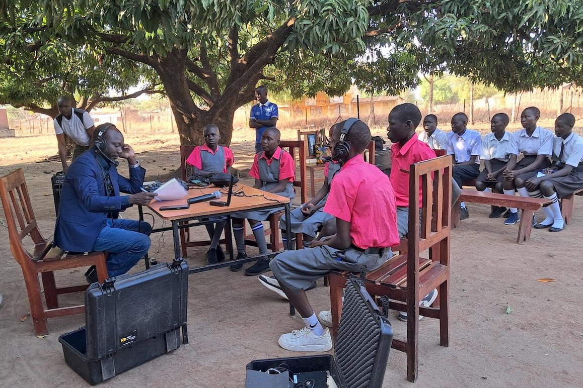 A man with a suit sits across a table with children in red t-shirts. All of the people at the table are wearing headphones and are recording a radio show. In the background, there are a group of students wearing blue shirts sitting watching the group at the table..