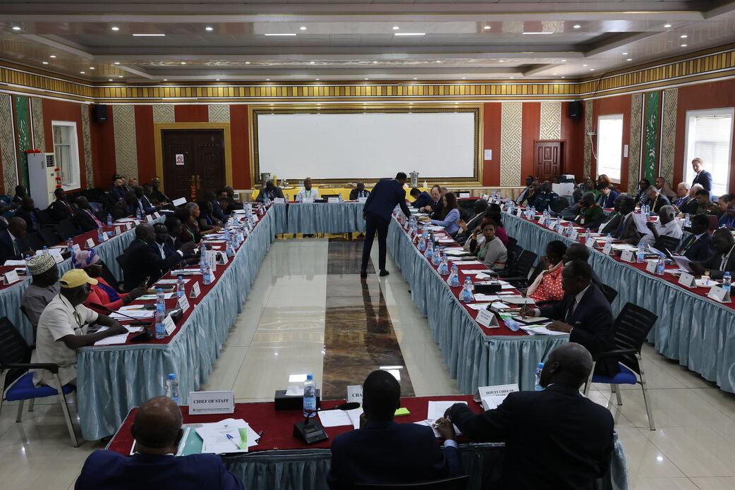 Large conference room with participants seated around a U-shaped arrangement of tables, facing a speaker at the center, with nameplates and documents on the tables.