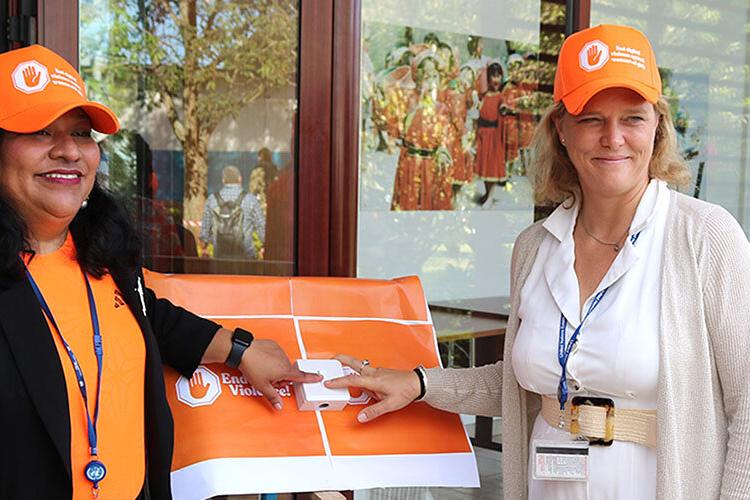 Two women both wearing orange coloured clothing and smiling each putting a finger on a button between them